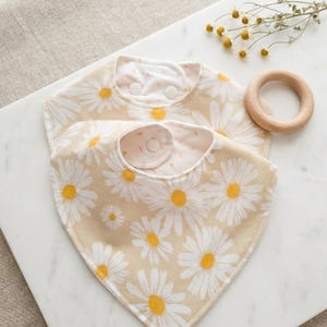 May include: Two daisy-patterned baby bibs with a beige background and white and yellow flowers. The bibs have a snap closure and are made of soft fabric. A wooden teething ring and dried flowers are also in the image.