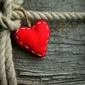 May include: A red heart-shaped ornament with white stitching hangs from a natural rope against a weathered wooden background. The heart is a symbol of love and affection, perfect for Valentine's Day or other special occasions.