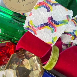 Op de afbeelding: Assortiment van kleurrijke lintrollen, waaronder een wit lint met regenboogpailletten, een groen fluwelen lint, een rood fluwelen lint, een groen lint en een blauw lint. De rollen zijn gelabeld "d. stevens fine ribbons".