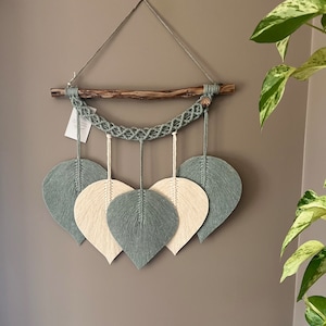 May include: A macrame wall hanging featuring a wooden branch and five leaf shapes. The leaves are crafted from yarn in shades of sage green and cream.