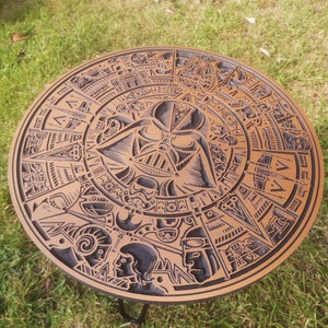 May include: A round wooden table top with a carved design featuring Darth Vader's face and other Star Wars characters. The table top is brown with black carvings.