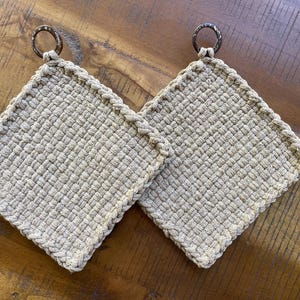 May include: Two beige woven square potholders with a braided edge and a circular metal ring for hanging. The potholders are made of a textured material and are displayed on a wooden surface.