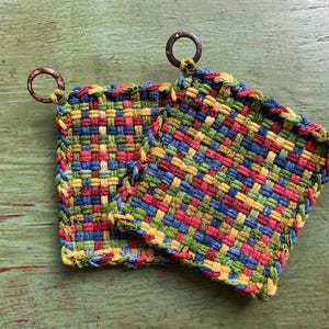 May include: Two colorful woven potholders with copper-colored hanging loops. The potholders feature a vibrant pattern of red, blue, green, and yellow threads. The potholders are square-shaped and appear handmade, perfect for kitchen use.