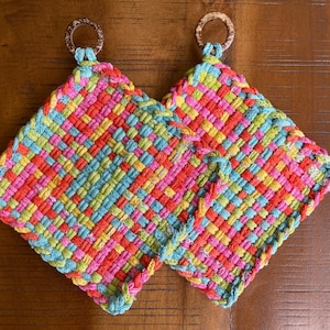 May include: Two colorful woven potholders with a square shape and a loop for hanging. The potholders are made of woven fabric in a vibrant pattern of red, pink, yellow, and blue. Each potholder has a wooden ring at the top.