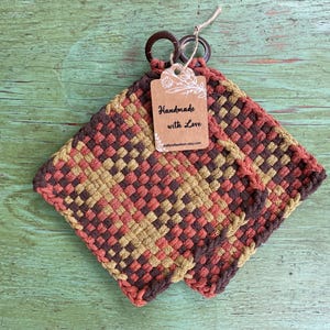 May include: Two woven square pot holders in shades of brown, peach, and olive green. Each pot holder has a dark brown loop for hanging. A tag reads "Handmade with Love".