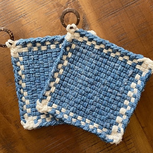 May include: Two square, woven potholders in shades of blue and white. The potholders have a woven texture with a white border and a loop for hanging. The potholders are made of a sturdy material and are suitable for kitchen use.