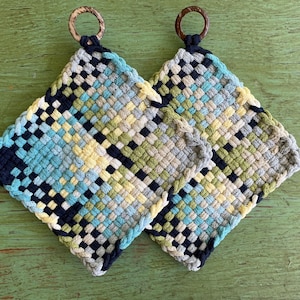 May include: Two square woven potholders with a colorful plaid pattern in shades of blue, green, yellow, and navy. Each potholder has a wooden ring for hanging. The potholders are displayed on a green wooden surface.