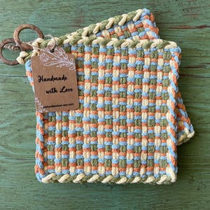 May include: Two woven square pot holders in shades of orange, blue, green, and cream. The pot holders have a woven design and a cream-colored border. A tag with the words "Handmade with Love" is attached with wooden rings.