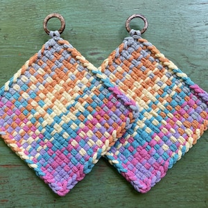 May include: Two colorful woven potholders with wooden ring handles. The potholders feature a diamond pattern in shades of orange, yellow, blue, pink, and purple. The potholders are square and appear to be handmade.