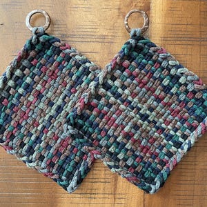 May include: Two woven potholders with a mix of colors including green, burgundy, and brown. Each potholder has a wooden ring for hanging. The potholders are square and have a braided edge.