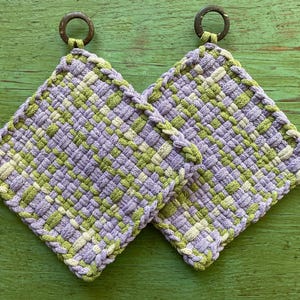 May include: Two square woven potholders in a lavender, green, and cream pattern. Each potholder has a woven loop for hanging and a braided edge. The background is a green wooden surface.