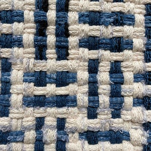 Puede incluir: Primer plano de un textil tejido con un patr&oacute;n repetitivo. El dise&ntilde;o presenta una cuadr&iacute;cula de cuadrados azules y blanquecinos alternados, creando una superficie texturizada. El tejido es apretado, con hilos visibles y ligeras variaciones de color.