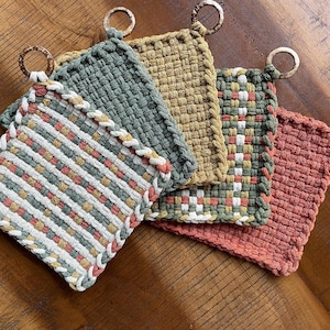 May include: A collection of five woven pot holders in various colors, including green, yellow, and coral. Each square pot holder has a loop for hanging and is made of a textured material. The pot holders are arranged on a wooden surface.