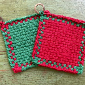 May include: Two woven potholders. One is green with red accents, the other is red with green accents. Both have a square shape and a loop made of wood for hanging. The potholders are made of a thick, textured material.
