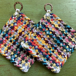 May include: Two colorful woven potholders with a variety of colors including red, blue, yellow, and green. Each potholder has a white border and a brown ring for hanging. The potholders are square-shaped and appear handmade.