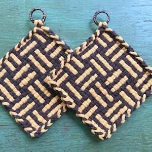 May include: Two square, woven potholders with a geometric pattern in navy blue and tan. Each potholder has a metal ring for hanging. The potholders are displayed on a teal wooden surface.