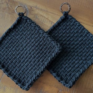 May include: Two dark gray woven potholders with wooden ring handles. The potholders are square-shaped with a textured surface and a braided edge. They are displayed on a wooden surface.