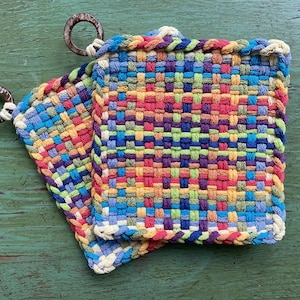 May include: Two colorful woven potholders with a square shape and a loop for hanging. The potholders feature a vibrant mix of red, blue, green, and yellow threads, creating a textured, handmade appearance. The background is a distressed green wooden surface.