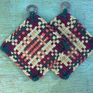 May include: Two woven potholders with a geometric pattern in burgundy, peach, cream, and teal. Each potholder has a loop for hanging. The potholders are square and made of a woven material.