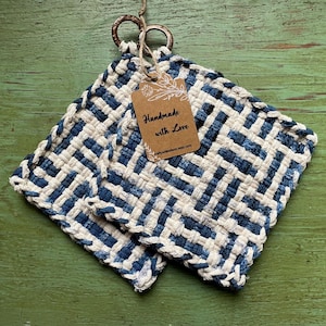 May include: Two square, woven potholders in blue and white, with a tag that reads "Handmade with Love." The potholders are made of a thick, textured material and have metal ring loops for hanging. The background is a distressed green surface.
