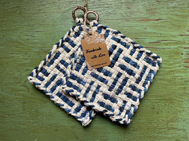 Puede incluir: Dos salvamanteles cuadrados, tejidos en azul y blanco, con una etiqueta que dice "Handmade with Love". Los salvamanteles est&aacute;n hechos de un material grueso y texturizado y tienen aros met&aacute;licos para colgar. El fondo es una superficie verde desgastada.