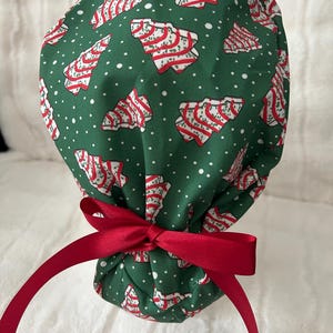 May include: A green scrub cap featuring a repeating pattern of white and red striped Christmas tree cookies and small white dots. The cap is gathered at the back and secured with a tied dark red ribbon bow at the base of the head.