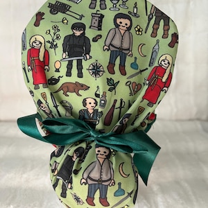 May include: A green scrub cap with a repeating pattern of medieval-themed cartoon figures and objects. The cap is tied with a dark green satin bow. The pattern includes figures in red and gray clothing, swords, and other medieval elements.