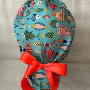 May include: A teal scrub cap featuring a colorful fish pattern in various sizes and designs. The cap is tied with a bright orange satin ribbon bow. The fish are in shades of pink, yellow, blue, and green.