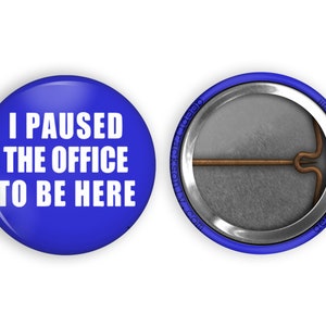 May include: A blue button with white text that reads "I paused the office to be here". The back of the button is silver with a pin.