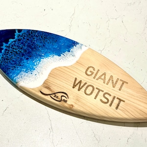 May include: A wooden surfboard serving tray with a blue and white epoxy resin wave design. The tray is engraved with the text "Giant Wotsit" and the logo "Fox Sea".