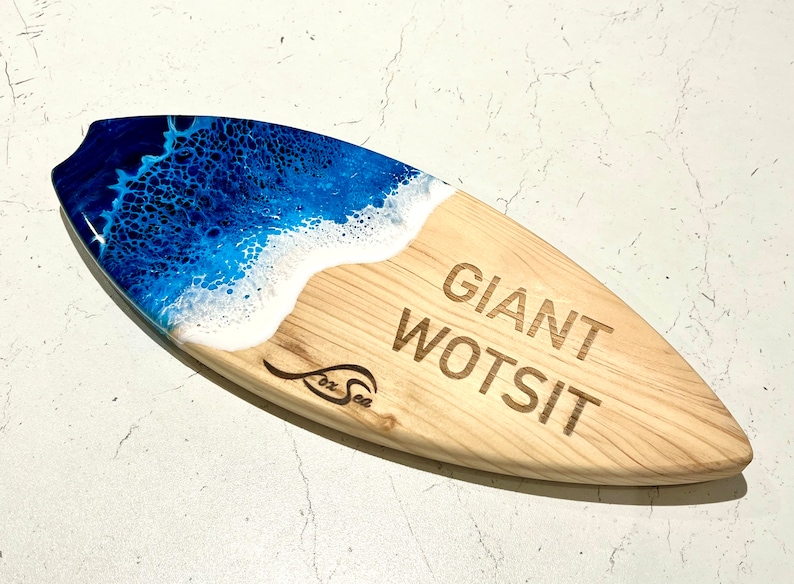 May include: A wooden surfboard serving tray with a blue and white epoxy resin wave design. The tray is engraved with the text "Giant Wotsit" and the logo "Fox Sea".