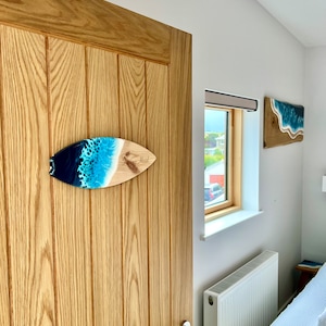 May include: A wooden surfboard with a blue and white resin ocean wave design. The surfboard is hanging on a wooden door with the word "Blood" written on it.