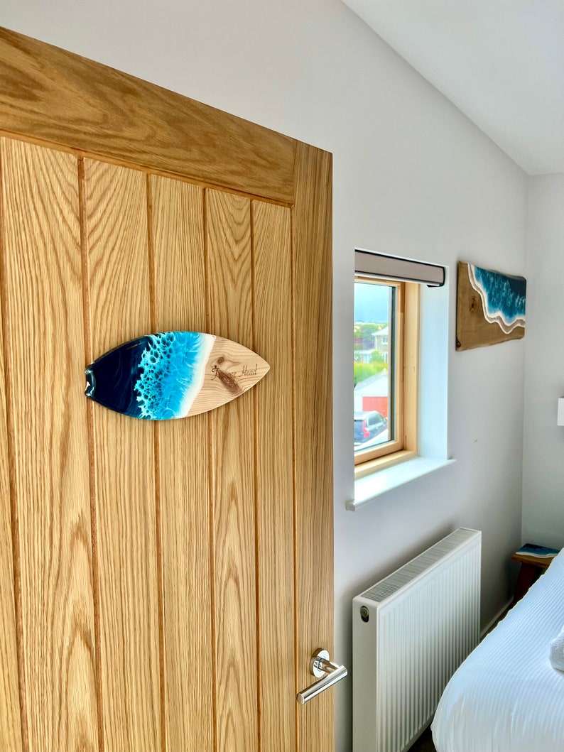 May include: A wooden surfboard with a blue and white resin ocean wave design. The surfboard is hanging on a wooden door with the word "Blood" written on it.