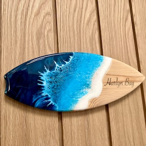 May include: A wooden surfboard with a blue and white resin ocean wave design. The surfboard is engraved with the words "Harlyn Bay".