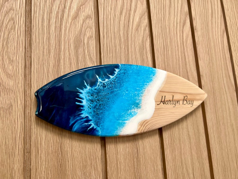 May include: A wooden surfboard with a blue and white resin ocean wave design. The surfboard is engraved with the words "Harlyn Bay".