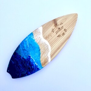 May include: A wooden surfboard with a blue and white resin ocean wave design. The surfboard has the dates "20.08.22", "22.03.22", and "20.08.22" engraved on it.