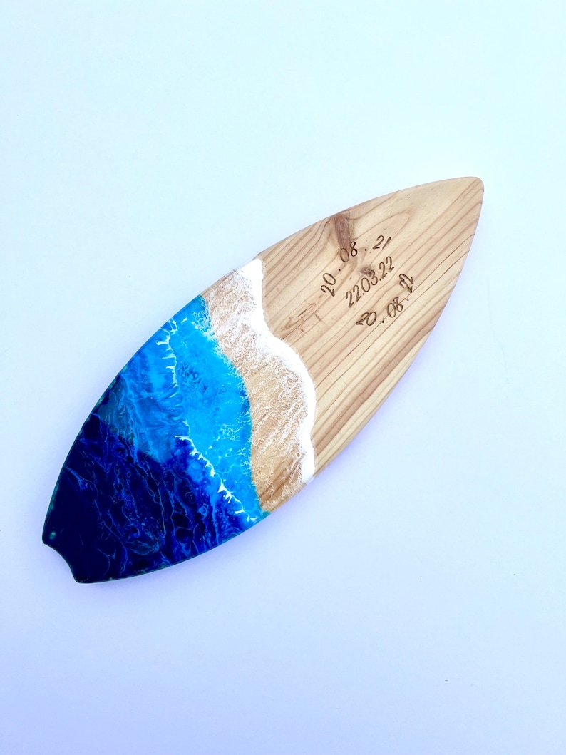 May include: A wooden surfboard with a blue and white resin ocean wave design. The surfboard has the dates "20.08.22", "22.03.22", and "20.08.22" engraved on it.