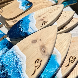 May include: A stack of wooden cutting boards with a blue and white ocean wave design. The boards are shaped like surfboards and have the words "Fox Sea" burned into the wood. Some of the boards have the name "Croyde" burned into the wood.