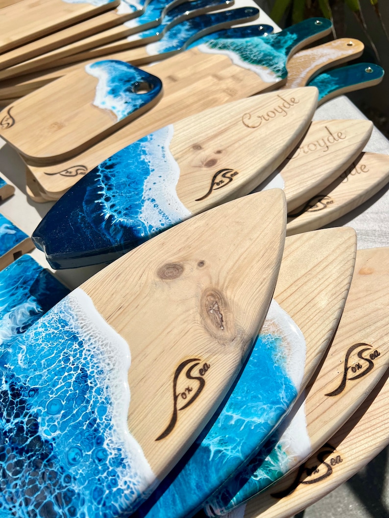 May include: A stack of wooden cutting boards with a blue and white ocean wave design. The boards are shaped like surfboards and have the words "Fox Sea" burned into the wood. Some of the boards have the name "Croyde" burned into the wood.