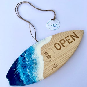May include: A wooden surfboard sign with a blue and white resin wave design. The sign says "OPEN" and has the text "The Bay Surf Shop" on it.