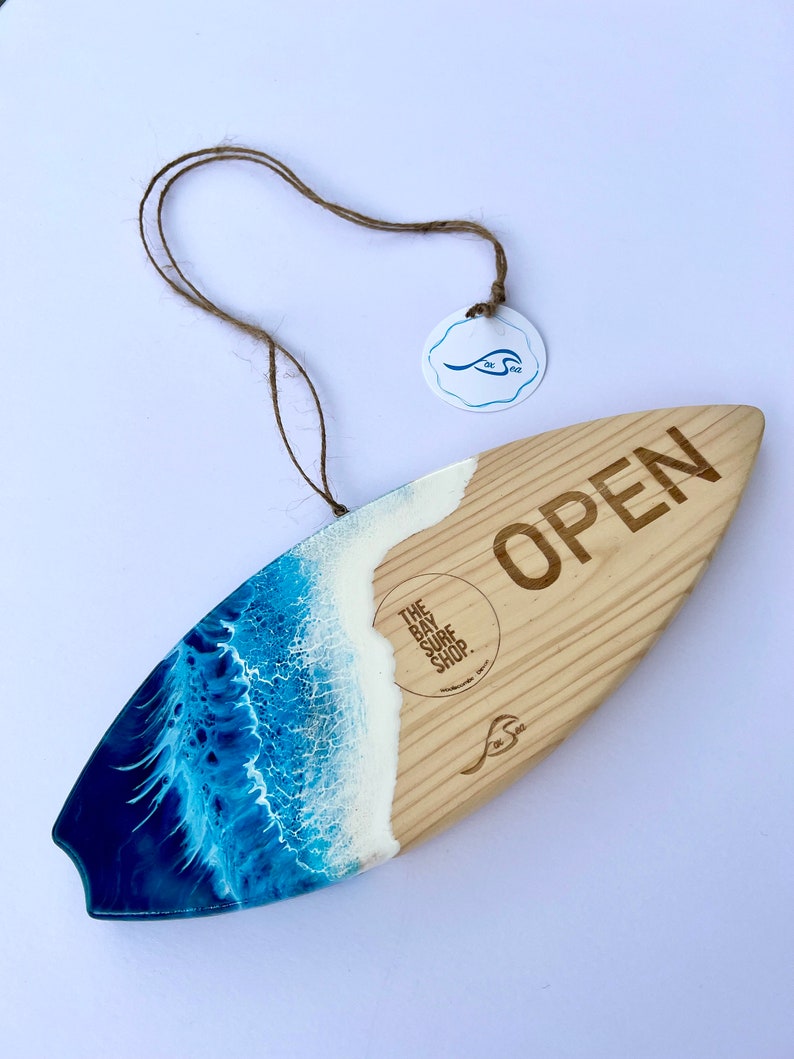 May include: A wooden surfboard sign with a blue and white resin wave design. The sign says "OPEN" and has the text "The Bay Surf Shop" on it.