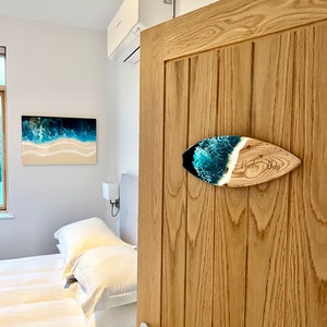 May include: A wooden surfboard sign with a blue and green resin inlay. The sign is mounted on a wooden door and says "Booby's Bay".