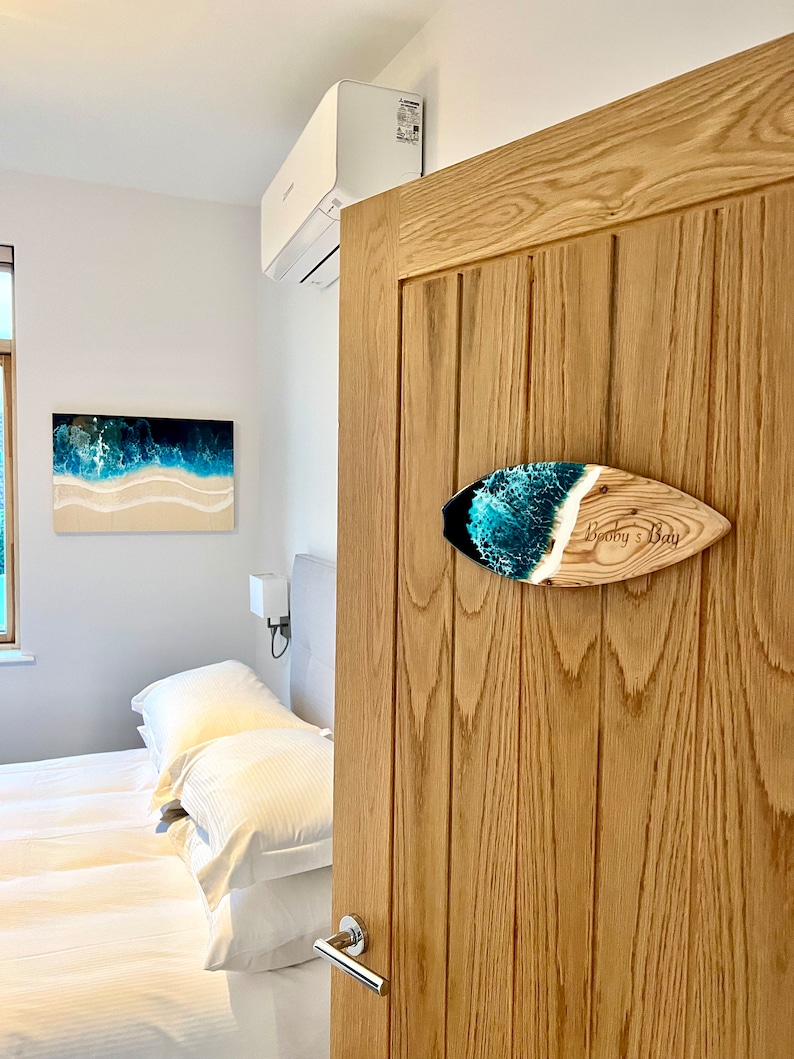 May include: A wooden surfboard sign with a blue and green resin inlay. The sign is mounted on a wooden door and says "Booby's Bay".
