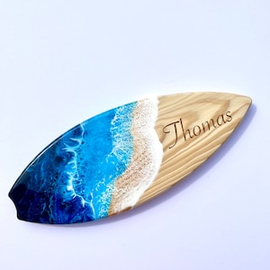 May include: A wooden surfboard with a blue and white resin ocean wave design. The surfboard is personalized with the name "Thomas" engraved on the wood.