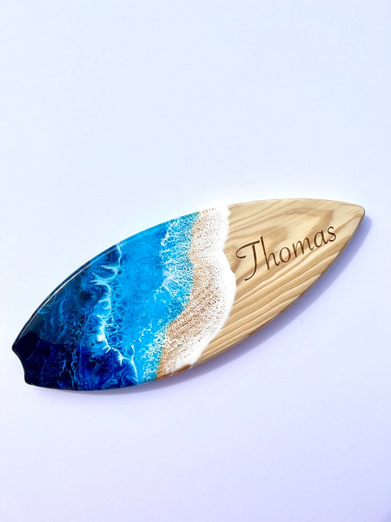 May include: A wooden surfboard with a blue and white resin ocean wave design. The surfboard is personalized with the name "Thomas" engraved on the wood.
