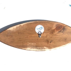 May include: A wooden surfboard wall decor with a distressed brown finish. The surfboard has a small metal hook on the back for hanging. The surfboard has a white sticker with the text "The Sea" and a wave design.