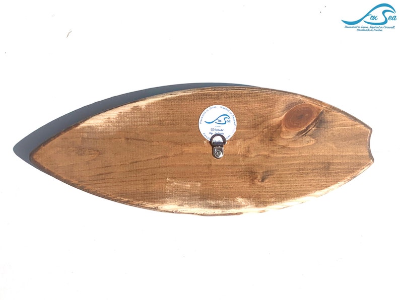 May include: A wooden surfboard wall decor with a distressed brown finish. The surfboard has a small metal hook on the back for hanging. The surfboard has a white sticker with the text "The Sea" and a wave design.