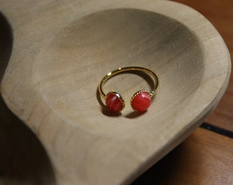 May include: A gold-colored open ring featuring two round, red-orange stones with white marbling. The ring is set in a simple, modern style, resting on a light brown wooden surface. The ring's design is minimalist and elegant.