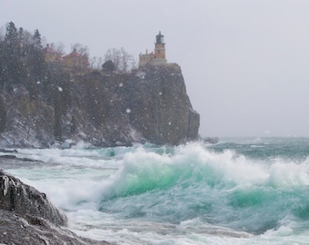 May include: A lighthouse perched on a rocky cliff overlooking a stormy sea with large waves crashing against the shore. Snow is falling and the water is a vibrant turquoise color.