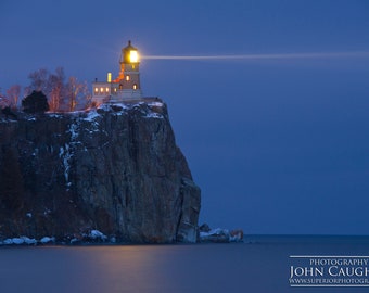 May include: A lighthouse on a cliff overlooking a dark blue body of water. The lighthouse is lit up and its beam shines across the water. The sky is a deep blue and the water is calm.